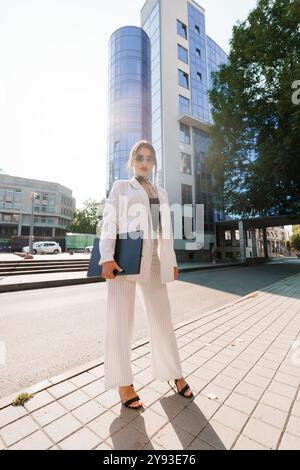 Selbstbewusste Geschäftsfrau im stilvollen Nadelstreifenanzug, die in urbaner Umgebung unter Sonnenlicht posiert. Stockfoto