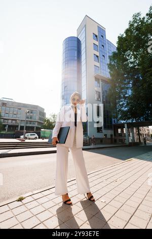 Stilvolle Geschäftsfrau im Nadelstreifenanzug mit Laptop gegen moderne Stadtarchitektur. Stockfoto