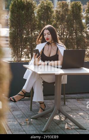 Elegante Geschäftsfrau in schicker Kleidung, die mit Laptop und Smartphone im Freien arbeitet. Stockfoto