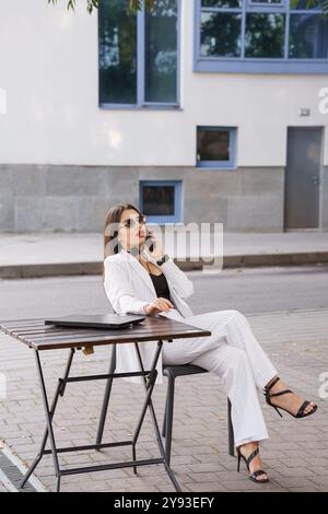 Stilvolle Geschäftsfrau im gestreiften Anzug, die am Tisch im Freien mit Laptop und Telefon sitzt. Stockfoto