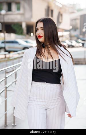 Stilvolle junge Frau mit langen Haaren, die einen trendigen Nadelstreifenanzug und roten Lippenstift trägt und selbstbewusst in einer urbanen Umgebung läuft. Stockfoto