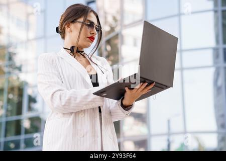 Stilvolle Geschäftsfrau in einem gestreiften Blazer, die in einem urbanen Ambiente mit ihrem Laptop interagiert. Stockfoto