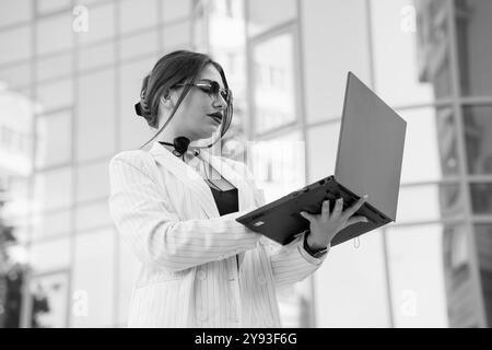 Stilvolle Geschäftsfrau im Nadelstreifenanzug, die mit Laptop außerhalb des modernen Gebäudes zusammenarbeitet. Stockfoto