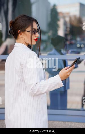 Stilvolle Geschäftsfrau im Nadelstreifenanzug, die sich mit dem Smartphone in urbaner Umgebung einlässt. Stockfoto