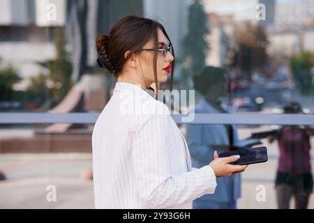 Professionelle Geschäftsfrau im stilvollen Nadelstreifenanzug, der das Smartphone hält, das die Stadtlandschaft reflektiert. Stockfoto