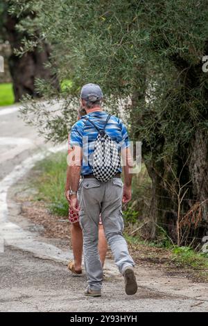 Ältere Ehepaare, die Rucksäcke oder Rucksäcke tragen, gehen gemeinsam auf einer ruhigen Straße auf der griechischen Insel Zante oder Zakynthos Stockfoto