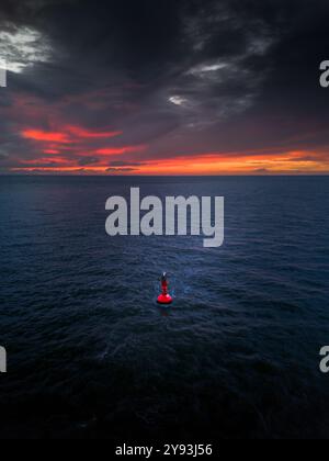 Eine einsame rote Boje schwingt im riesigen Ozean unter einem dramatischen Sonnenuntergangshimmel, mit dunklen Wolken und leuchtenden Rottönen, die den Horizont beleuchten. Stockfoto