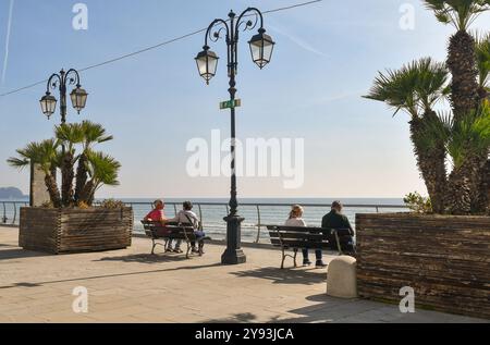 Zwei Paare mittleren Alters sitzen auf den Bänken der Promenade und genießen die Frühlingssonne, Alassio, Savona, Ligurien, Italien Stockfoto