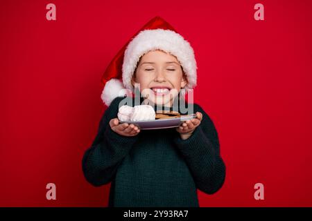 Foto-Porträt des charmanten kleinen Mädchens riechen leckere Snacks gekleidet grüne weihnachtskleidung feiern Winterferien auf rotem Hintergrund Stockfoto