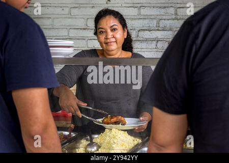 Eine fröhliche Verkäuferin serviert ein traditionelles El-Salvadorianisches Gericht von ihrem Street Food-Stand zu einem Gast. Die Umgebung zeigt ein lässiges, lebhaftes Ambiente Stockfoto