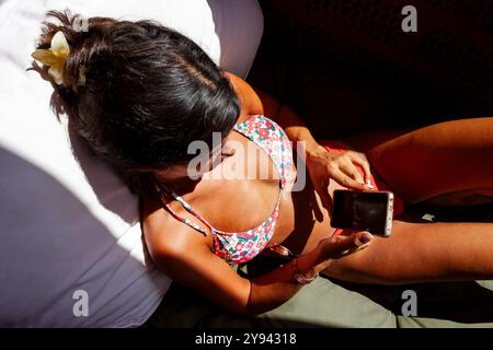 Eine Frau in einem farbenfrohen Bikini wird von oben erfasst, während sie ihr Smartphone benutzt und das Sonnenlicht in Barcelona am frühen Morgen genießt. Sie schaut nach unten Stockfoto