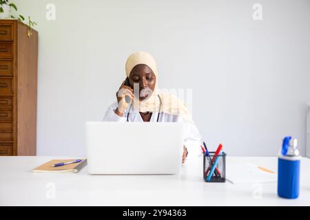Eine professionelle schwarze Ärztin mit einem Hijab ist in einem Telefongespräch beschäftigt, während sie ihren Laptop in einer hellen medizinischen Praxis verwendet Stockfoto
