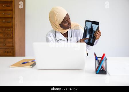 Eine professionelle schwarze Ärztin mit Hijab untersucht Röntgenbilder auf einem Tablet und sitzt an ihrem Schreibtisch in einem modernen klinischen Büro Stockfoto