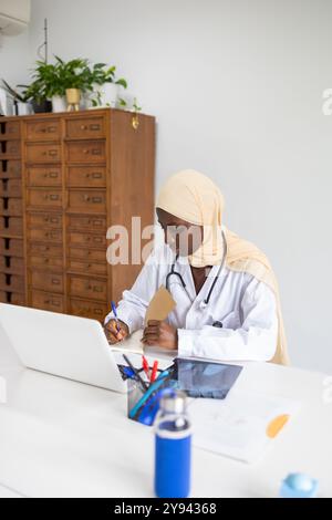 Eine professionelle schwarze Ärztin mit Hijab konzentriert sich darauf, Notizen zu schreiben, während sie an ihrem Schreibtisch in einer gut beleuchteten, modernen Büroumgebung sitzt, Ref Stockfoto