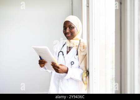 Professionelle schwarze Ärztin, die einen Hijab und einen Laborkittel trägt, eine medizinische Akte hält und in einem gut beleuchteten Raum an einem Fenster steht und einen Konkurrenten reflektiert Stockfoto