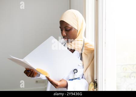 Eine schwarze Ärztin, die einen Hijab und ein Stethoskop trägt, konzentriert sich sehr darauf, ein Dokument zu lesen, während sie in der Nähe eines Fensters in ihrem Büro steht Stockfoto