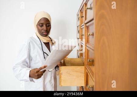 Eine professionelle afrikanische Ärztin, die einen Hijab trägt, berät Dokumente, während sie neben einer offenen Holzschublade steht Stockfoto
