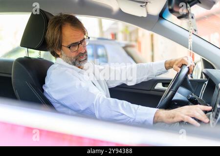 Ein reifer Geschäftsmann in einem weißen Hemd passt die Bedienelemente in seinem Auto an, während er nach unten blickt und sich auf seine Fahraufgaben konzentriert. Stockfoto