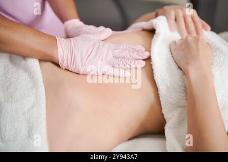 Die abgeschnittene, nicht erkennbare professionelle Masseurin trägt pinkfarbene Handschuhe und bietet einer auf einem Therapientisch liegenden Patientin eine wohltuende Bauchmassage. T Stockfoto