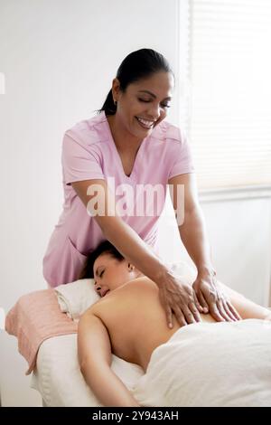 Eine Frau erhält eine entspannende Rückenmassage von einer lächelnden Therapeutin in einem hellen Zuhause. Die Empfängerin hat ihre Augen geschlossen und genießt den Ter Stockfoto