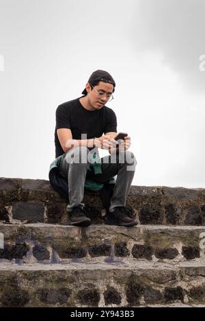 Ein junger mexikanischer Wanderer hält an einer Steinmauer inne, um sein Smartphone zu benutzen. Er trägt eine schwarze Mütze, ein T-Shirt und eine Brille, draußen mit bewölktem Himmel Stockfoto