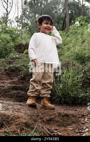 Ein kleiner Junge lächelt hell, während er auf einem Waldweg steht, in lässiger Wanderkleidung. Sein fröhlicher Ausdruck und die natürliche Kulisse vermitteln ein Gefühl von A Stockfoto