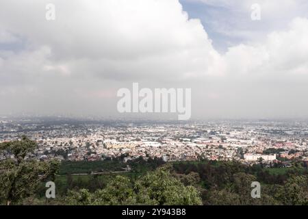 Verbesserte Aussicht von einem Hügel aus auf die riesige, geschäftige Landschaft einer mexikanischen Stadt, umgeben von üppigem Grün, unter einem bewölkten Himmel Stockfoto