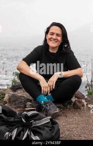 Ein fröhlicher mexikanischer Wanderer sitzt während einer Pause auf einem felsigen Felsvorsprung und überblickt eine riesige, in Nebel gehüllte Stadtlandschaft. Sie ist in Schwarz gekleidet und mit lebhaftem blau Stockfoto