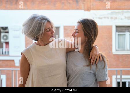Ein herzerwärmendes Porträt einer Mutter mit silbernem Haar und ihrer Tochter, die sich auf einem Balkon liebevoll umarmen, beide lächeln sanft und strahlen Wärme aus Stockfoto