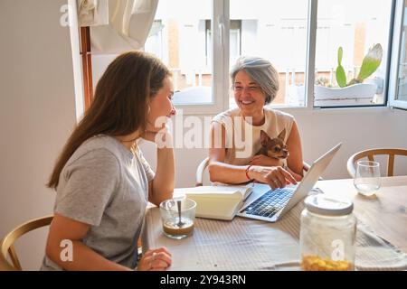 Eine fröhliche Mutter und Tochter sitzen an einem Küchentisch mit einem Laptop und teilen einen leichten Moment zusammen in einem hellen, heimeligen Setting Einen kleinen Hund und öffnen n Stockfoto