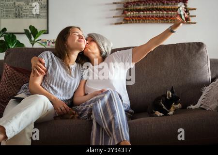 Eine Mutter mittleren Alters und ihre Tochter im Teenageralter teilen sich einen liebevollen Kuss, während sie ein Selfie auf einem bequemen Sofa machen. Kleine Hunde sitzen neben ihnen auf der Couch Stockfoto