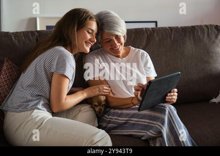 Eine fröhliche Mutter und Tochter teilen sich einen Moment der Bindung, während sie auf einem gemütlichen Sofa ein Tablet betrachten, in dem sich ein kleiner Hund zwischen ihnen schmiegt Stockfoto