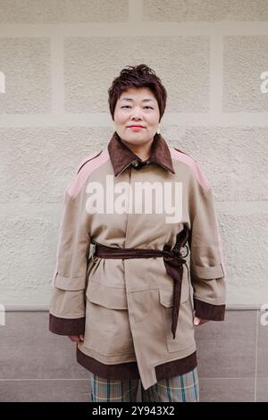 Eine asiatische Frau mittleren Alters steht vor einer beigefarbenen Wand, gekleidet in einem schicken Trenchcoat mit braunen Details, der Selbstvertrauen und Anmut ausstrahlt Stockfoto