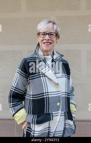 Eine fröhliche Seniorin mit blondem Haar und Brille, in einem modischen schwarz-weißen karierten Mantel, lächelt warm an einer weichen beigefarbenen Wand Stockfoto