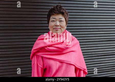 Eine reife asiatische Frau lächelt warm und trägt einen auffälligen rosa Schal vor einem strukturierten dunklen Hintergrund, die mit ihrem kurzen Haar und ihren minimalistischen Ohrringen zusammengehalten wird Stockfoto