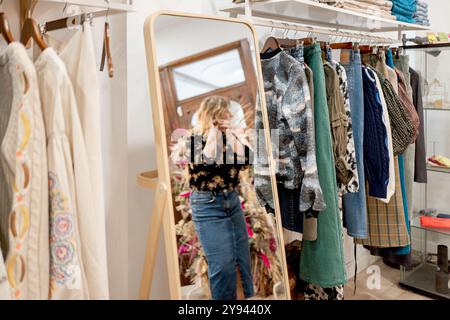 Eine Frau steht vor einem großen Spiegel in einer eleganten Boutique. Die Boutique bietet Regale mit modischen Jacken und Kleidung, die einen Welcomi widerspiegeln Stockfoto