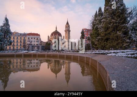 Winter in Madrid, wo Sie eine Kirche und moderne Gebäude, die mit Schnee bedeckt sind und sich wunderschön in einem nahegelegenen Teich unter dem Dämmerungshimmel spiegeln Stockfoto