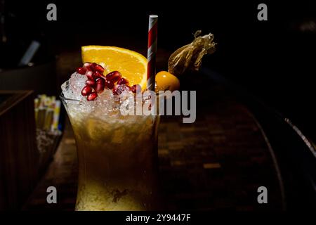 Ein erfrischender Fruchtcocktail garniert mit Orangenscheibe, Granatapfelkernen und Physalis, serviert in einem hohen Glas mit Eis, serviert in einem dimmeren Glas Stockfoto