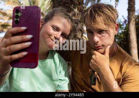 Eine junge Frau und ein Mann genießen die gemeinsame Zeit am Strand, während die Frau ein Selfie mit ihrem Smartphone macht, während der Mann nachdenklich neben ihr posiert Stockfoto