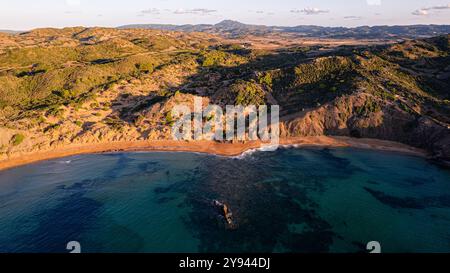 Eine atemberaubende Drohnenaufnahme des sonnendurchfluteten Cavalleria Beach auf Menorca mit dem lebendigen blauen Wasser, der zerklüfteten Küste und der üppigen grünen Umgebung Stockfoto