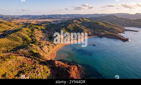 Atemberaubende Drohnenaufnahme des Cavalleria Beach auf Menorca mit dem goldenen Sonnenuntergang, das die zerklüftete Küste und das klare blaue Wasser umrahmt Stockfoto