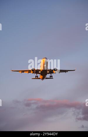 Ein kommerzielles Flugzeug steigt durch einen ruhigen Himmel hinab, mit einem orangefarbenen Sonnenuntergang, der eine leuchtende Kulisse für seinen glatten Flug darstellt Stockfoto