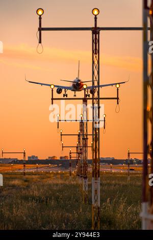 Ein kommerzielles Flugzeug bereitet sich auf die Landung vor, während es zwischen beleuchteten Lichtern vor einem lebendigen Sonnenuntergang absteigt Stockfoto