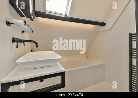 Ein elegantes und modernes Bad mit eckigem Waschbecken, mattschwarzen Armaturen und einer schrägen Decke mit Fenster. Stockfoto