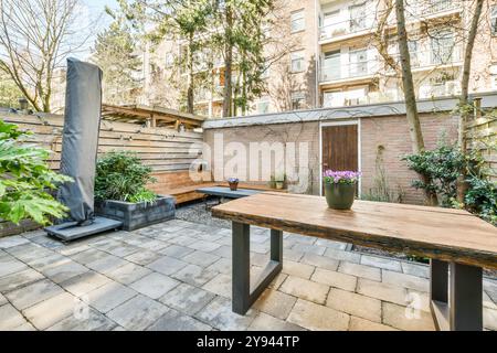 Ein ruhiger urbaner Garten mit Holztisch, Bänken und lebendigen Pflanztöpfen vor einem Hintergrund von üppigem Grün und einem Wohngebäude. Stockfoto