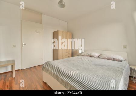 Ein gut beleuchtetes Schlafzimmer mit minimalistischem Design, einem Doppelbett mit grauer Bettwäsche, einem Holzschrank und einer einfachen Bank. Stockfoto