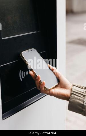 Eine Nahaufnahme einer Hand, die ein Smartphone in einer Autoladestation hält, eine Frau, die kontaktlos bezahlt, um ihr Auto für Elektrofahrzeuge aufzuladen. Stockfoto