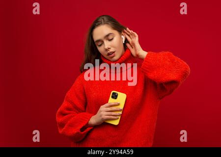 Schöne emotionale junge Frau, die Musik in schnurlosen Ohrstöpseln mit einem modernen Smartphone in einem Studio hört, mit rotem Hintergrund Stockfoto