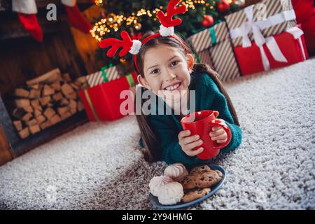 Großes Foto des niedlichen kleinen Mädchens liegend Boden Snacks Kakao gekleidet grün weihnachten Outfit Tageslicht Haus Party Dekoration Innenraum Zimmer Stockfoto