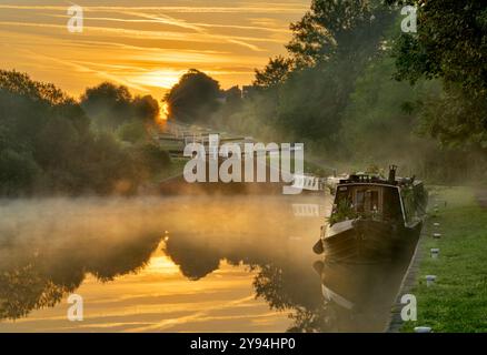 Caen Hill schließt bei Sonnenaufgang, in Devizes mit niedrig liegendem Nebel und einem wunderschönen warmen Glühen und Kanalbooten vor Anker Stockfoto